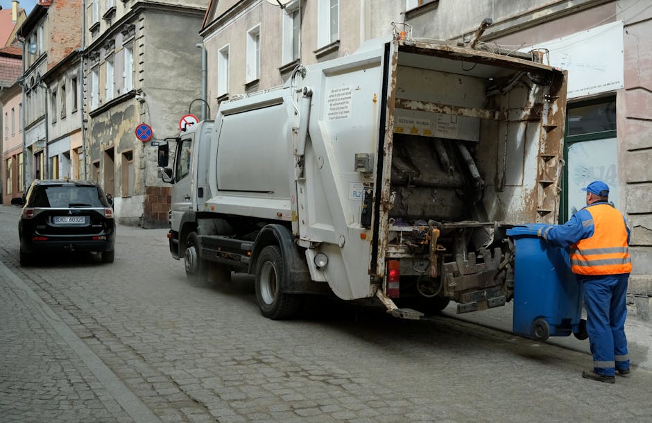 A sanitation worker dressed in a high-visibility orange vest and blue uniform is disposing of waste into a large white rubbish collection vehicle parked on a cobblestone street in an urban area. The truck's rear compartment is open, revealing rusted metal components and a hydraulic mechanism used for compacting rubbish. The worker is holding a blue wheeled bin, which he is tilting to deposit waste into the vehicle. To the left, a black car with visible license plates is parked parallel to the curb, and the surrounding buildings feature aged facades with multiple windows and signage. The street is narrow, lined with multi-storey structures exhibiting a mix of stone and plaster finishes, with some peeling paint and weathered surfaces. The scene appears to be during daytime, with natural light illuminating the environment, emphasizing the practicality of onsite waste collection as an alternative to other disposal methods. The environment subtly highlights the importance of professional rubbish removal services like those provided by companies such as rubbishclearancekentishtown.co.uk for private waste management needs.