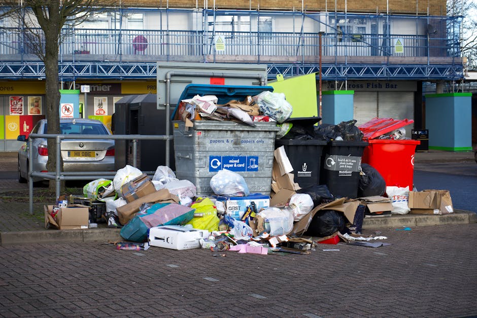 An outdoor scene in front of a commercial building shows a large accumulation of overflowing rubbish and waste materials on a paved pavement area. The waste includes numerous cardboard boxes, some flattened and others still containing items, mixed paper and cardboard packaging, black and green plastic rubbish bags, and discarded packaging debris. At the centre, a large grey recycling bin labeled for mixed paper and card is open with waste spilling out, surrounded by smaller black, red, and black composite bins. Behind the rubbish, there is a silver car parked parallel to the waste pile, with a visible European-style yellow license plate. The background features a multi-storey building with a blue scaffolding and protective railings, suggesting ongoing construction or maintenance work. On either side of the waste, there are two vertical green and blue frames, possibly part of waste disposal or recycling facilities. The environment appears to be an urban or suburban shopping area, with storefronts partially visible in the distance, emphasizing the context of private waste collection or rubbish removal services such as those provided by rubbishclearancekentishtown.co.uk. The lighting suggests daytime with natural light, and the scene portrays the typical need for efficient rubbish removal outside commercial premises.