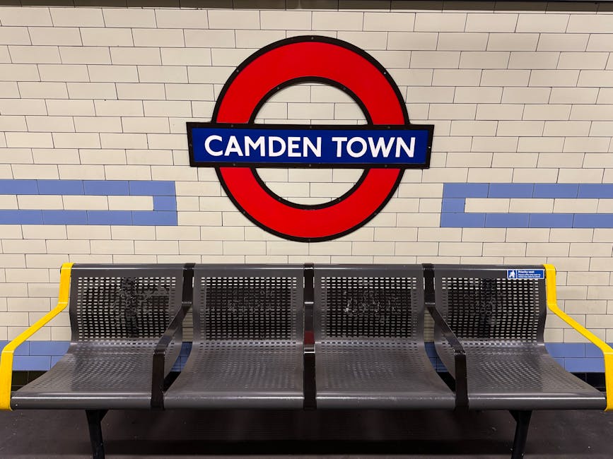 A row of four metal benches with perforated surfaces and black frames, positioned along a tiled wall inside Camden Town underground station. The wall is finished with white bricks and features a large London Underground roundel sign with a red outer circle, black border, and a blue rectangular panel in the center displaying white text reading 'CAMDEN TOWN.' To the right of the benches, a small blue and white sign indicates priority seating. The benches have bright yellow armrests on both ends, providing safety and visibility. The overall environment appears clean and well-maintained, with natural lighting illuminating the area, reflecting typical urban public transport surroundings. The scene subtly supports the concept of public spaces where waste management or rubbish clearance services may be relevant when considering station upkeep or station-based waste collection options.