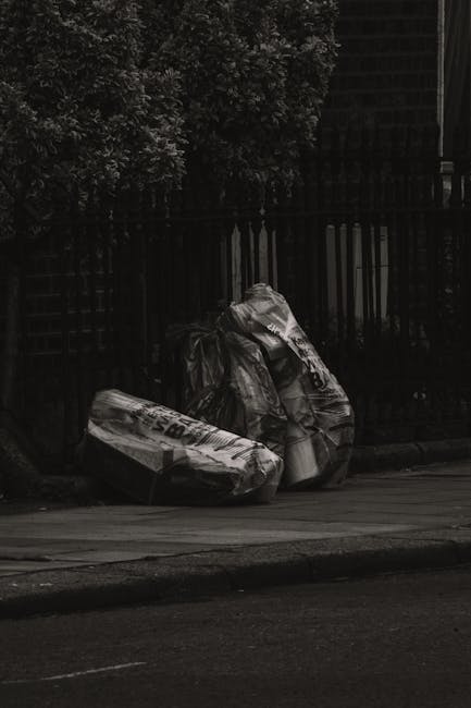 Three large black plastic garbage bags filled with rubbish are placed on a grey sidewalk near a black metal fence, which runs parallel to the curb. The bags are crumpled and tied at the top, with some sections appearing shiny and wrinkled, indicating plastic material. Behind the fence, dense foliage of dark leaves is visible, and to the right, part of a building with dark bricks and windows can be seen. The scene is set on a quiet street with a paved road in the foreground, and the lighting suggests it is either early morning or late evening, with muted illumination. This image, sourced from rubbishclearancekentishtown.co.uk, illustrates a typical outdoor scene where waste is temporarily stored prior to collection or disposal in line with alternative private rubbish removal methods common in residential areas of Kentish Town, NW5.
