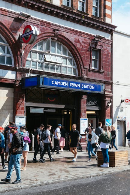 The entrance of Camden Town underground station featuring a large blue sign with white text reading 'CAMDEN TOWN STATION' mounted above the doorway, with a red-brick architectural facade. Several pedestrians, dressed in casual clothing and carrying backpacks or bags, are walking in and out of the station on a paved sidewalk. Some individuals appear to be waiting or conversing nearby. The station's entrance is flanked by windows with curved, white-framed panes, and an underground roundel sign is mounted on the brickwork above the entrance. The environment suggests a busy urban setting typical for a well-frequented transit hub, with surrounding buildings visible and a partly cloudy sky overhead. The scene subtly relates to urban waste management by showing an active public space where rubbish removal and street sanitation services may operate to maintain cleanliness around high-footfall areas, such as Camden Town.