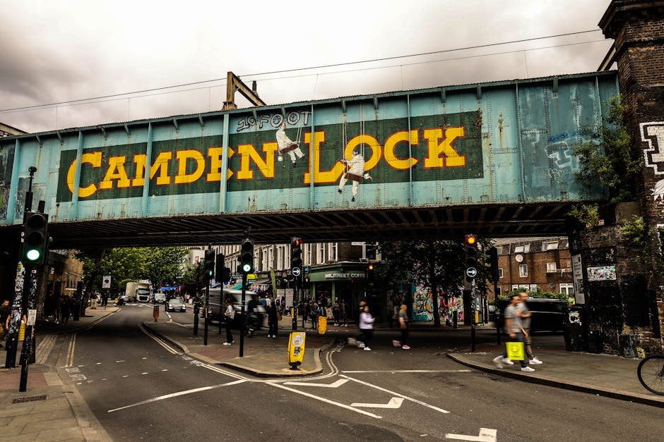 A large, weathered steel railway bridge painted in a faded turquoise color spans across a busy urban street in Camden, with the words ' Camden Lock' prominently displayed in bold yellow letters on the side of the bridge. The surface of the bridge shows signs of rust and peeling paint, indicating age and exposure to the elements. Two small murals of children climbing the bridge are visible, painted in a simple, cartoonish style. Below the bridge, the street scene includes pedestrians crossing at the crosswalk, some walking briskly and others standing near the traffic signals, which are illuminated with green lights. On the road, there are visible lane markings, with a few parked vehicles and a bus in the distance. The environment suggests a typical day in a vibrant, bustling neighborhood, with the bridge serving as an iconic feature that could be associated with independent or private waste handling, including rubbish removal services available in the area. Tall brick buildings and a mix of urban greenery surround the scene, with some graffiti and street art visible on nearby walls, adding to the lively atmosphere. The lighting suggests an overcast day, contributing to the subdued yet dynamic cityscape depiction.