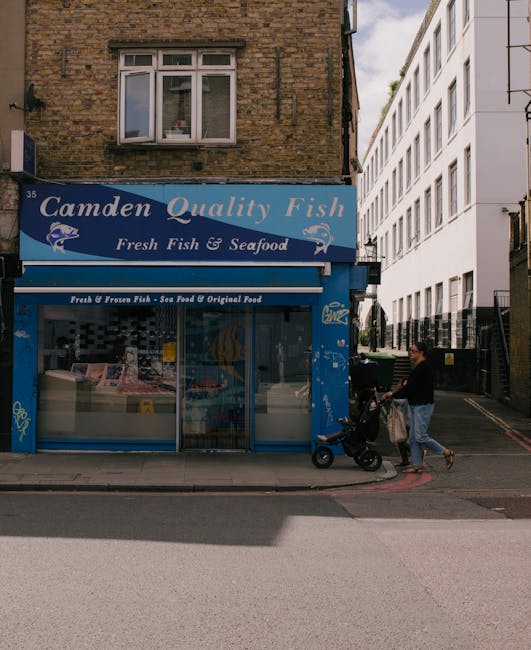 The entrance of Camden Town underground station featuring a large blue sign with white text reading 'CAMDEN TOWN STATION' mounted above the doorway, with a red-brick architectural facade. Several pedestrians, dressed in casual clothing and carrying backpacks or bags, are walking in and out of the station on a paved sidewalk. Some individuals appear to be waiting or conversing nearby. The station's entrance is flanked by windows with curved, white-framed panes, and an underground roundel sign is mounted on the brickwork above the entrance. The environment suggests a busy urban setting typical for a well-frequented transit hub, with surrounding buildings visible and a partly cloudy sky overhead. The scene subtly relates to urban waste management by showing an active public space where rubbish removal and street sanitation services may operate to maintain cleanliness around high-footfall areas, such as Camden Town.