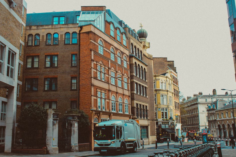The image depicts a daytime urban street scene with a row of multi-story buildings featuring varied architectural styles, including brick facades, ornate window frames, and decorative top sections. In the foreground, a white waste collection vehicle is parked on the pavement, partially obscuring the entrance to what appears to be a private property fenced by white stone pillars and wrought iron gates. The vehicle is positioned on the right side of the street, with its front facing towards the camera, indicating active rubbish collection or on-site clearance. Surrounding the vehicle are cobblestone or paved surfaces with small bollards mitigating vehicle access. To the left, part of a modern building with large windows and a beige exterior contrasts with the older, more ornate structures. In the background, more historic and contemporary buildings line the street, with features such as domed roofs and decorative cornices. The scene is illuminated by natural daylight, contributing to a clear and neutral atmosphere suitable for illustrating private waste handling and alternative rubbish removal services in an urban setting, supporting SEO relevance for rubbish clearance and disposal in a city environment.