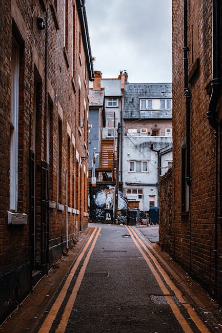 A narrow urban alleyway flanked by tall brick buildings on both sides, with a paved surface featuring yellow double lines along the edges, leading towards a back area in the distance. The scene includes a variety of waste containers and debris at the end of the alley, adjacent to the buildings, indicating ongoing rubbish clearance or disposal activities. The background shows a mix of aged, weathered facades with several small windows and a metal staircase attached to one of the buildings, suggesting a utilitarian space likely used for service access or private waste handling outside typical public rubbish collection routes. Overcast sky above provides diffuse lighting, emphasizing the textured brickwork and concrete surfaces. Such an environment may require independent rubbish removal services to manage accumulated waste efficiently in this densely built, private urban setting, aligning with alternative waste management practices associated with private or on-site clearance solutions.