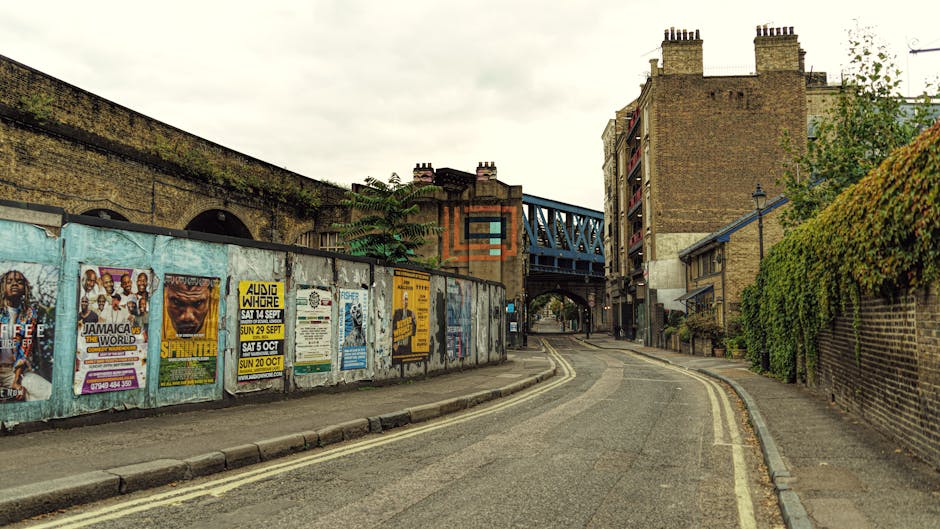 A narrow urban street scene showing an upward-curving road with a worn asphalt surface, marked by double yellow lines along one side. On the left, a weathered blue-painted concrete wall is covered with various posters advertising events, including reggae concerts, DJ nights, and cultural festivals, some partially torn or faded. The wall extends towards a bridge with a blue metal structure supporting railway tracks that cross overhead. On the right, a brick wall with lush green ivy growing over its surface borders a sidewalk, which is paved with rectangular concrete slabs. Behind the wall, multi-storey residential buildings with brick facades and multiple balconies are visible, featuring chimneys extending from the rooftops. The scene is lit by diffuse daylight under an overcast sky, creating a subdued, neutral atmosphere typical of British urban streets. The overall aesthetic highlights an area that may be subject to independent waste collection or private rubbish clearance services, with no visible rubbish or waste, just typical street elements and signage.