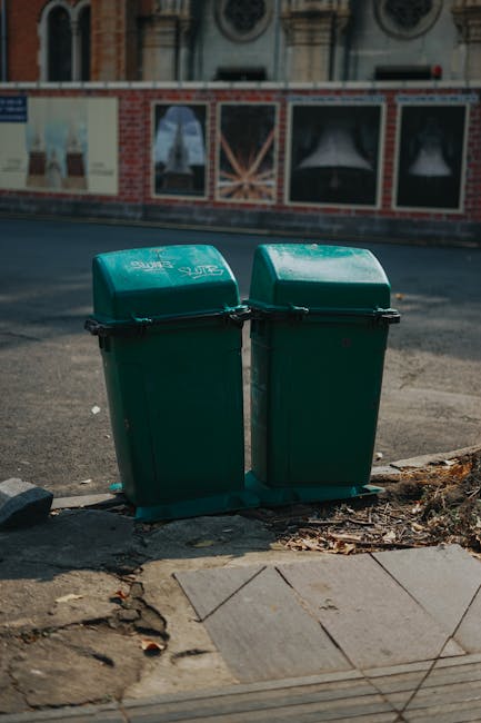 Two green plastic rubbish bins with hinged lids are positioned side by side on a concrete sidewalk at the edge of a street in an urban area. The bins have a textured surface and appear to be made of durable polypropylene with a matte finish. They are placed close to a small curb, with scattered debris and dried leaves around their base. In the background, there is a brick wall with a decorative red and white patterned design, and a row of windows partially visible above the wall. The street surface is asphalt, and a parked vehicle can be seen faintly in the distance. The lighting suggests daytime with natural light, creating soft shadows on the ground. The scene indicates an area where residents might independently handle rubbish disposal, supported by local waste clearance services such as those offered by Kentish Town waste management providers, aligning with the context of alternative rubbish collection and on-site waste handling.