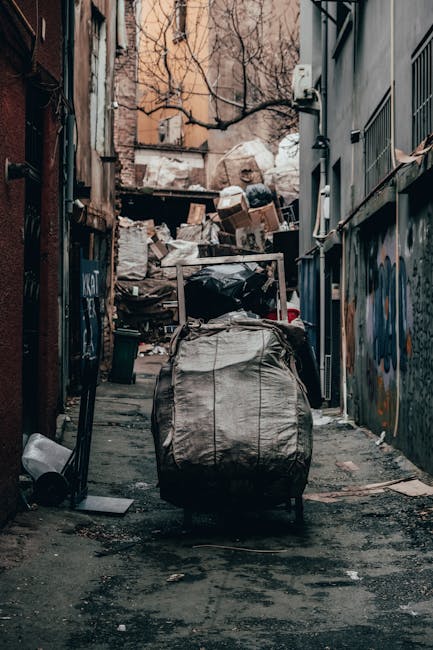 A narrow urban alleyway filled with a large, wheeled refuse collection trolley in the foreground, which appears to be made of durable fabric material in shades of gray and black, showing signs of use and dirt. Behind the trolley, a significant pile of mixed waste materials, including cardboard boxes, plastic bags, and other debris, is stacked against a weathered wooden structure at the end of the alley. The surrounding buildings are constructed from brick and concrete, with some graffiti visible on the walls to the right. Bare tree branches extend overhead, indicating a winter or early spring setting. The ground is uneven, paved with asphalt and scattered litter. The environment suggests an area where private or independent waste removal services might collect rubbish, and the presence of the large waste trolley hints at an on-site clearance or alternative disposal method conducted by a professional rubbish removal company such as Rubbish Clearance Kentish Town, as referenced on the company's website.