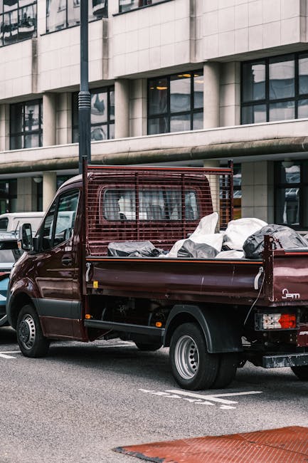 A small burgundy flatbed truck parked on a street in front of a modern office building with large glass windows. The truck's cargo area is filled with various debris and waste materials, including black garbage bags and unidentifiable rubbish. The vehicle has a protective metal grille behind the cab, and the bed is partially loaded, with some items leaning against the sides. The scene is set during daylight, with a streetlamp visible close to the vehicle, and other parked cars are seen in the background. This image illustrates a typical scene of private waste collection or rubbish clearance, as carried out by companies offering independent rubbish removal services in urban environments, highlighting the disposal of builders' waste or unwanted items from property clearance activities. The urban setting and the presence of waste materials relate to non-local authority rubbish disposal methods, exemplifying how such waste may be temporarily stored before collection or disposal at recycling centres or waste facilities.