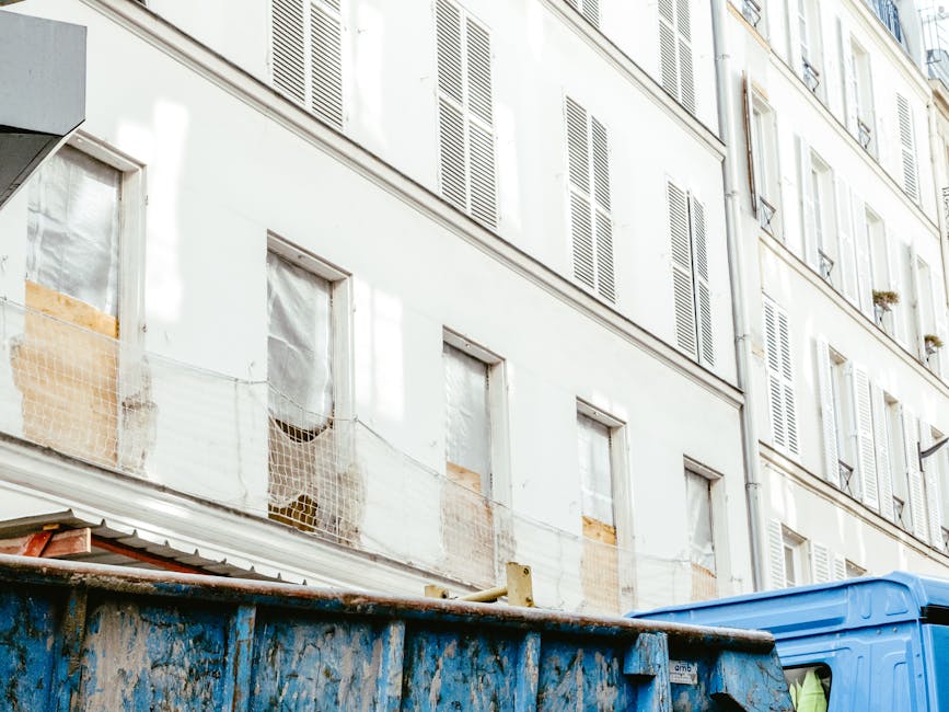 The image depicts the upper floors of a multi-storey residential building with a light-colored facade and multiple windows fitted with white shutters. Some windows are boarded up with wooden panels, and others are covered with transparent protective sheeting. A horizontal wire runs across the middle of the building, supporting a mesh safety net. In the foreground, a large, weathered blue skip with chipped paint and rust stains is positioned on a paved surface, partially blocking the view of the building's lower part. The skip contains various discarded materials and waste, suggesting ongoing rubbish clearance or renovation work. To the right, the corner of a blue van is visible, parked next to the skip, supporting the context of waste removal or private disposal services. The scene is naturally lit, capturing the textures of the building’s exterior, the metallic finish of the skip, and the overall environment, which emphasizes the typical setting for independent rubbish collection and waste management during property renovation or refurbishment.