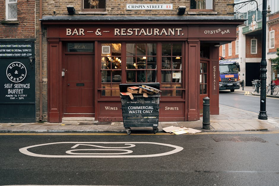 A black commercial waste bin labeled 'COMMERCIAL WASTE ONLY' is positioned on the sidewalk directly in front of a reddish-brown storefront that functions as a bar and restaurant. The bin is overflowing with flattened cardboard boxes and paper debris, some of which spill onto the pavement. To the left of the bin, there is a small black signboard advertising a self-service buffet, and to the right, a black bollard and a metal pole separate the sidewalk from the street. A large white pedestrian crossing symbol is painted on the asphalt road in the foreground. In the background, a colorful truck is parked on the street, and a few bicycles are secured to a bike rack near a multi-story building. The scene appears to be in an urban area with typical street furniture, and the waste bin’s placement suggests it may be an example of improper refuse disposal, which relates to private waste handling and rubbish removal considerations.